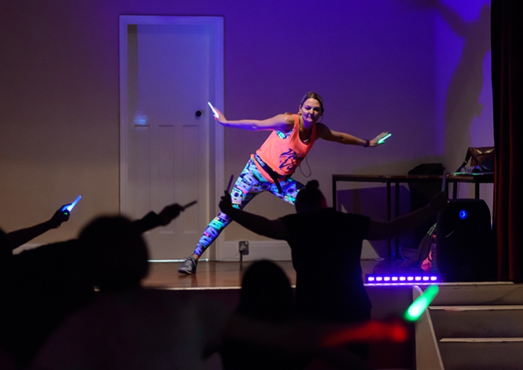 A woman on a stage peforms aerobics in the dark, with neon lights and glow sticks
